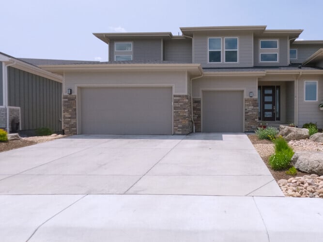 A contemporary two-story house features stone and wood siding with a landscaped yard, concrete driveway and a mix of plants and rocks in the front yard, which are highlighted under daylight.